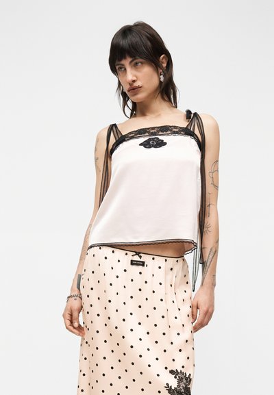 Γυναίκα με σκούρα μαλλιά, που φοράει λευκό σατέν καμησόλ με μαύρη δαντέλα και φούστα ανοιχτό ροζ με πουά και μαύρες ανθικές λεπτομέρειες.