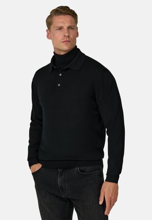 Uomo che indossa una polo nera a maniche lunghe sopra un dolcevita nero, con le mani nelle tasche di jeans scuri, guardando leggermente di lato.
