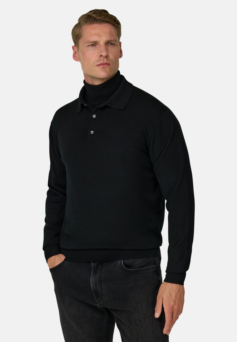 Homme portant un polo noir à manches longues par-dessus un col roulé noir, les mains dans les poches d'un jean foncé, regardant légèrement sur le côté.