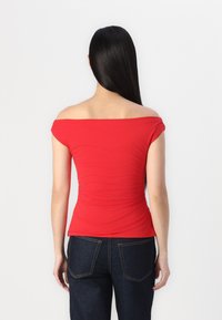 Red off-the-shoulder top with gathered texture at the back, paired with dark denim jeans featuring standard pockets and stitching accents.