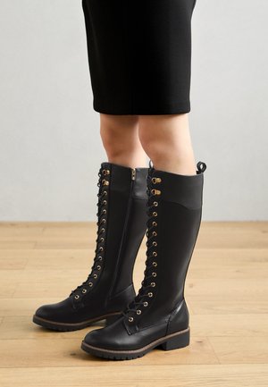 Črne usnjene škornje do kolen z zlatimi očesci in vezalkami. Imajo gladko teksturo, stransko zadrgo in nizko blokasto peto.