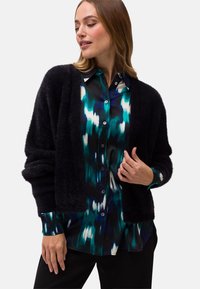 Svart fluffig cardigan med långa ärmar, bärs över en flerfärgad sidenblus med blå och gröna abstrakta mönster, knapprad stil.