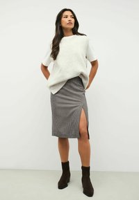 Vesta tricotată de culoare gri deschis peste un tricou alb, asortată cu o fustă cu carouri, ajustată, care are o fanta laterală și botine maro.