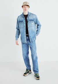 Camicia di jeans azzurro chiaro con due taschini sul petto, abbinata a jeans azzurro chiaro e sneakers multicolori. Il cappello aggiunge un accento neutro.