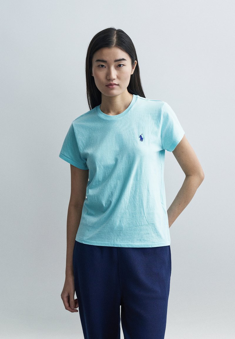 Gaiši zilā īsroku kokvilnas T-krekls ar apaļu kaklu un mazu izšūtu logotipu. Kombinēts ar tumši zilām biksēm uz neitrāla fona.