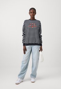 Maglione a righe blu navy e bianco con grafica rossa "NYC 1990", abbinato a jeans chiari a gamba larga e sneakers bianche, mentre tiene una borsa bianca.