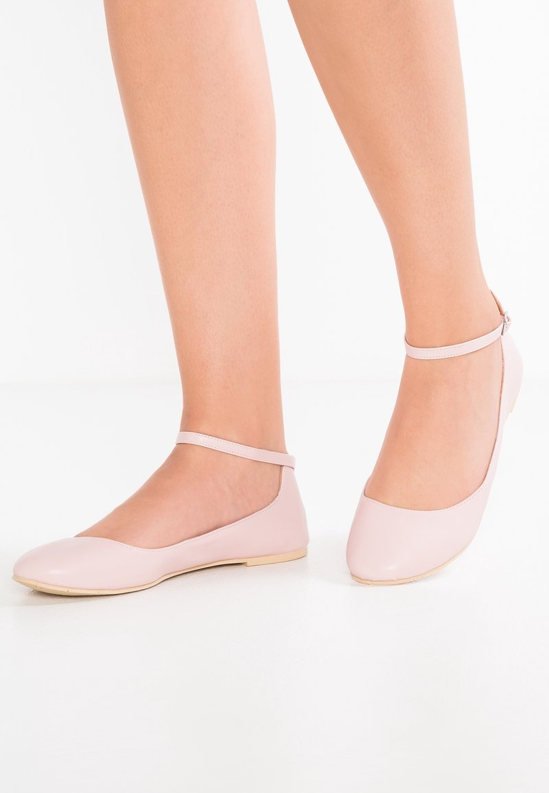 Jambes portant des chaussures plates rose clair avec des brides à la cheville sur une surface et un fond blancs.