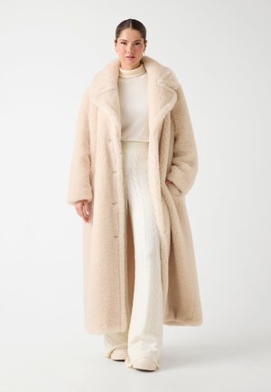 Femme portant un long manteau en fausse fourrure beige sur un pull col roulé crème et un pantalon en tricot blanc texturé, se tenant contre un fond uni.