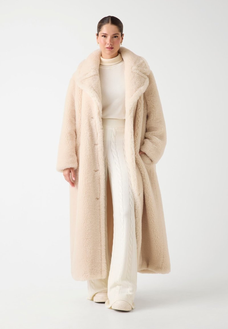 Donna che indossa un lungo cappotto in pelliccia sintetica beige sopra un maglione a collo alto crema e pantaloni bianchi lavorati a maglia, in piedi contro uno sfondo semplice.
