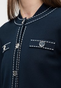 Κοντινό πλάνο ενός μπλε ναυτικού cardigan με λευκή ραφτή λεπτομέρεια, ασημένια κουμπιά και ψηλό γιακά που φοράει ένα άτομο.