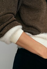 Pruunist lõngast kootud sviiter valgete kunstkarvadega mansettide ja ribakonstruktsiooniga. Suurendatud vaade säärele ja varruka detailile neutraalses taustas.