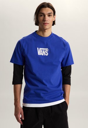 Junger Mann mit mittellangen dunklen Haaren, der ein blaues Vans-T-Shirt über einem schwarzen Langarmshirt trägt und vor einem einfachen hellen Hintergrund steht.