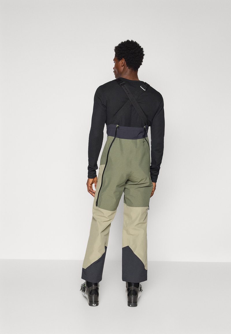 Kombinezon narciarski w kolorze zielonym i beżowym, z projektem pasa harness, kieszeniami zapinanymi na zamek i rozszerzonym krojem. Teksturowana tkanina z zestawieniem kolorów.