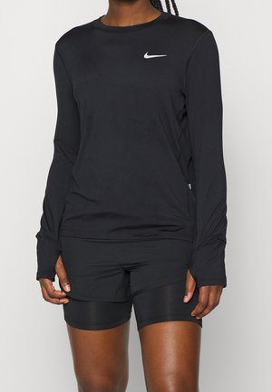 Osoba nosząca czarną, długorękawową sportową koszulkę Nike z otworami na kciuki i czarne dopasowane spodenki rowerowe na jednolitym tle.