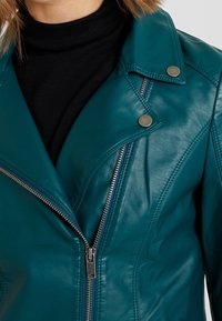 Turkusowa skórzana kurtka z ukośnym zapięciem na zamek, zatrzaskami i dopasowanym krojem. Gładka faktura i wyraźne detale szwów.