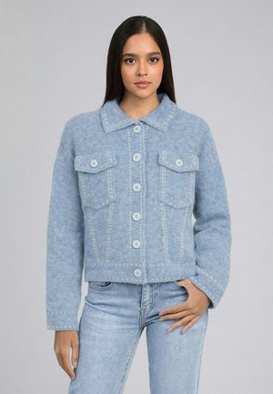 Jeune femme aux longs cheveux bruns portant une veste en laine bleu clair à boutons et un jean bleu clair, posant devant un fond gris uni.