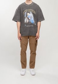 Grafické oversized tričko v vybledlém uhlíkovém odstínu s designem modrého anděla, spárované s khaki cargo kalhotami s několika kapsami a bílými teniskami.
