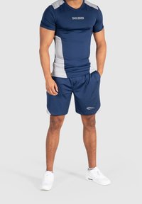 Camiseta atlética azul marino con acentos grises, de mangas cortas y un diseño ajustado, combinada con pantalones cortos deportivos azul marino que presentan un logo reflectante. Zapatillas blancas.