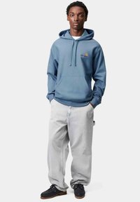 Sudadera de algodón azul con bolsillo frontal, capucha ajustable y logo. Combinada con unos pantalones baggy gris claro y zapatos negros, de pie sobre un fondo sencillo.