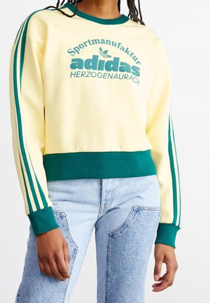 Personne portant un sweat Adidas crème et vert avec texte et bandes vertes sur les manches, associé à un jean bicolore bleu clair.