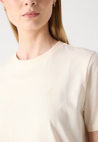 T-shirt in cotone beige con colletto rotondo, caratterizzata da un logo diamantato dorato discreto sul lato sinistro del petto. Tessuto morbido, maniche corte.