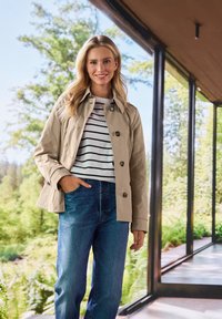 Beige Leichtjacke mit Kragen, Knopfverschluss und zwei Taschen. Getragen über einem gestreiften Hemd und kombiniert mit blauen Jeans.