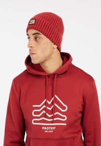 Rode gebreide beanie met ribbeltextuur en een genaaide label. Draagt met een bijpassende rode hoodie met een witte grafische print en trekkoorden.