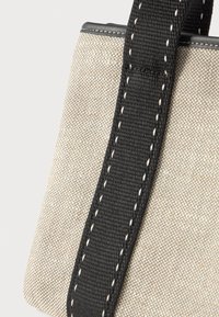 Κοντινό πλάνο από μπεζ υφασμένο τσαντάκι με ευρεία μαύρη λουρί που διαθέτει άσπρες ραφές κατά μήκος των άκρων.