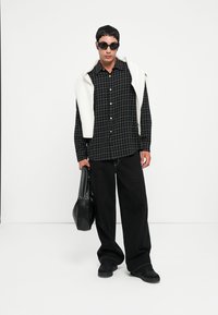 Czarna koszula w kratę z guzikami, oversize'owe czarne spodnie, czarne buty, dziergany kremowy sweter zarzucony na ramionach, oraz czarne okulary przeciwsłoneczne. W zestawie czarna torba.