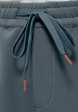 Cordón atado en un lazo en la cintura elástica azul grisácea de pantalones deportivos, mostrando costuras detalladas y tela texturizada en primer plano.