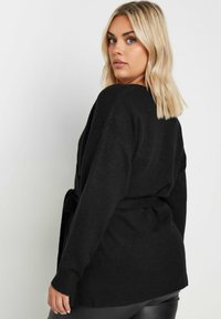 Svart, öppen cardigan i mjukt, texturerat stickat tyg. Har en bälte i midjan och löst sittande ärmar.