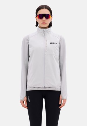 Woman wearing light gray zip-up vest over long-sleeve shirt, black leggings with white logo, and red reflective sports sunglasses, standing straight.