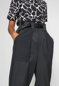 Svarta cargo-byxor med strukturerade lappar, elastisk midja och bälte. De har stora sidofickor och ett mjukt, lätt tyg.