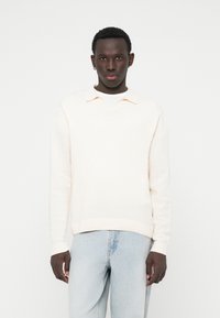 Lys cremefarvet strikket sweater med krave, der har en v-udskæring og ribbede manchetter, parret med lys blå denim jeans.