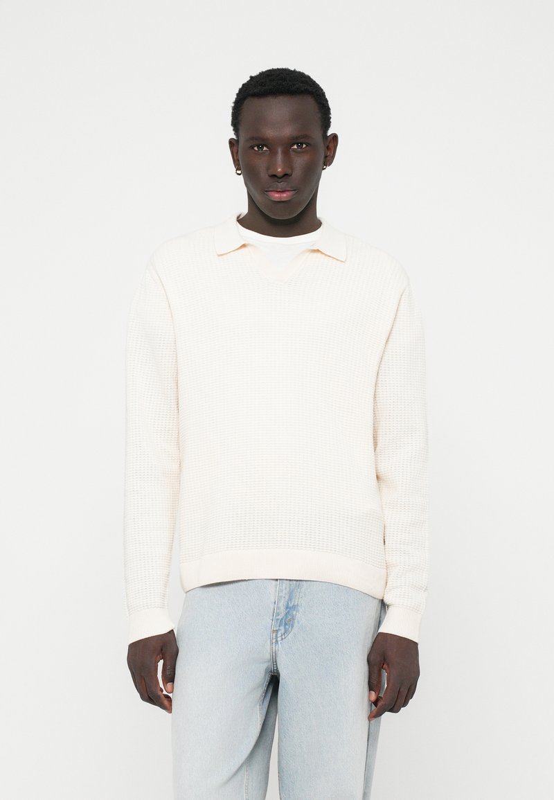 Lys cremefarvet strikket sweater med krave, der har en v-udskæring og ribbede manchetter, parret med lys blå denim jeans.