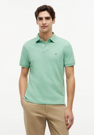 Ung mand iført en lysegrøn polo og beige bukser, stående med den ene hånd i lommen mod en ensfarvet hvid baggrund.