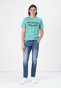 Light teal short-sleeve t-shirt with black “PME LEGEND” print, paired with faded blue jeans and white sneakers, standing against a white background.