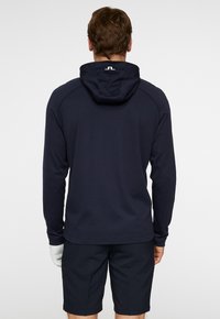 Mörkblå pullover hoodie med en figurnära design, texturerad tyg och huva. Har en logotyp på baksidan av halsringningen. Kombineras med mörka shorts.
