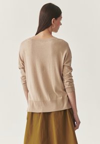 Beige kötött pulóver laza szabással, bordás mandzsettákkal és szegéllyel. A háta enyhén hosszabb, és széles nyakkivágással rendelkezik. Olíva zöld szoknyával párosítva.