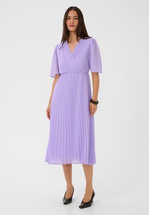 Femme portant une robe midi plissée violet clair avec des manches volantées et des talons noirs, debout devant un fond blanc uni.