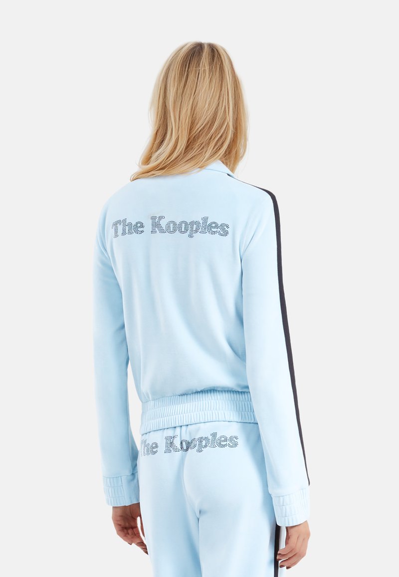 Lichtblauwe velours trainingsjas met "The Kooples" in strass-steentjes, zwarte zijstrepen en geribde manchetten en zoom.