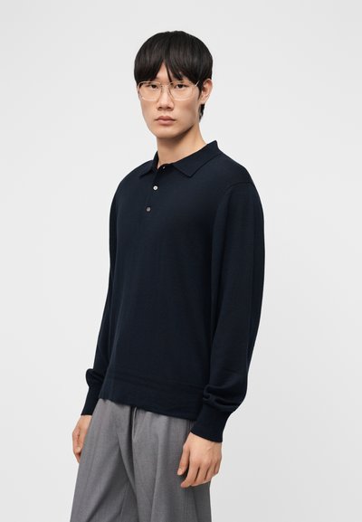 Noor mees lühikeste mustade juuste ja prillidega, kellel on seljas tumesinine pika varrukaga polo-särk ja heledat halli värvi püksid, taust on ühevärviline.