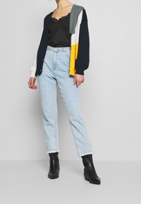 Cardigan a maglia multicolore con sezioni blu navy, grigio, bianco e giallo, indossato su un top in pizzo nero e jeans a vita alta blu chiaro.