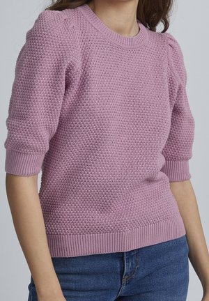 Frau trägt einen strukturierten, hellvioletten Kurzpullover mit Puffärmeln, kombiniert mit blauen Jeans.