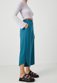 Tyrkysové culottes s bočními kapsami a vysokým pasem, doplněné o krémový top s krátkým rukávem, který má žebrovanou texturu a zvonové rukávy. Černé ploché sandály.