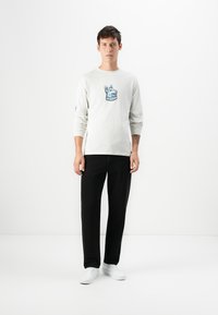 Lange mouwen lichtgrijze t-shirt met een blauwe hondenafbeelding op de borst, gecombineerd met zwarte broek en witte sneakers, staande tegen een effen achtergrond.