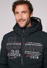 Giacca a vento nera con zip frontale, realizzata in tessuto texture, con cappuccio e loghi ricamati bianchi/rossi e coordinate sul petto.