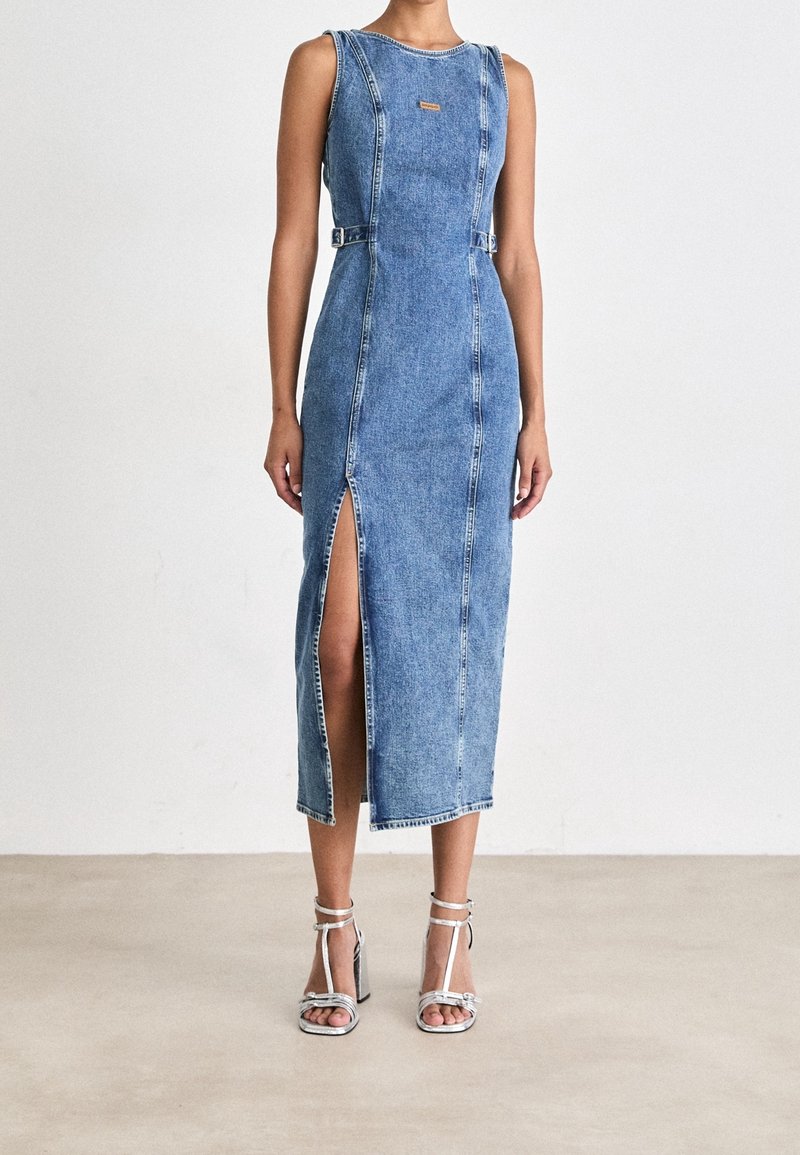 Femme portant une robe midi en denim bleu sans manches avec une fente devant et des détails de boucles à la taille, associée à des sandales blanches à talons hauts.