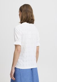Hvid bluse i øjelet design med korte volantsleeves; har broderi og et struktureret mønster overalt. Bæres med en blå nederdel.