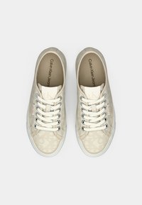 Sneaker bassa beige di Calvin Klein con lacci bianchi, occhielli in metallo e motivi logo sottili, viste dall'alto su uno sfondo bianco.